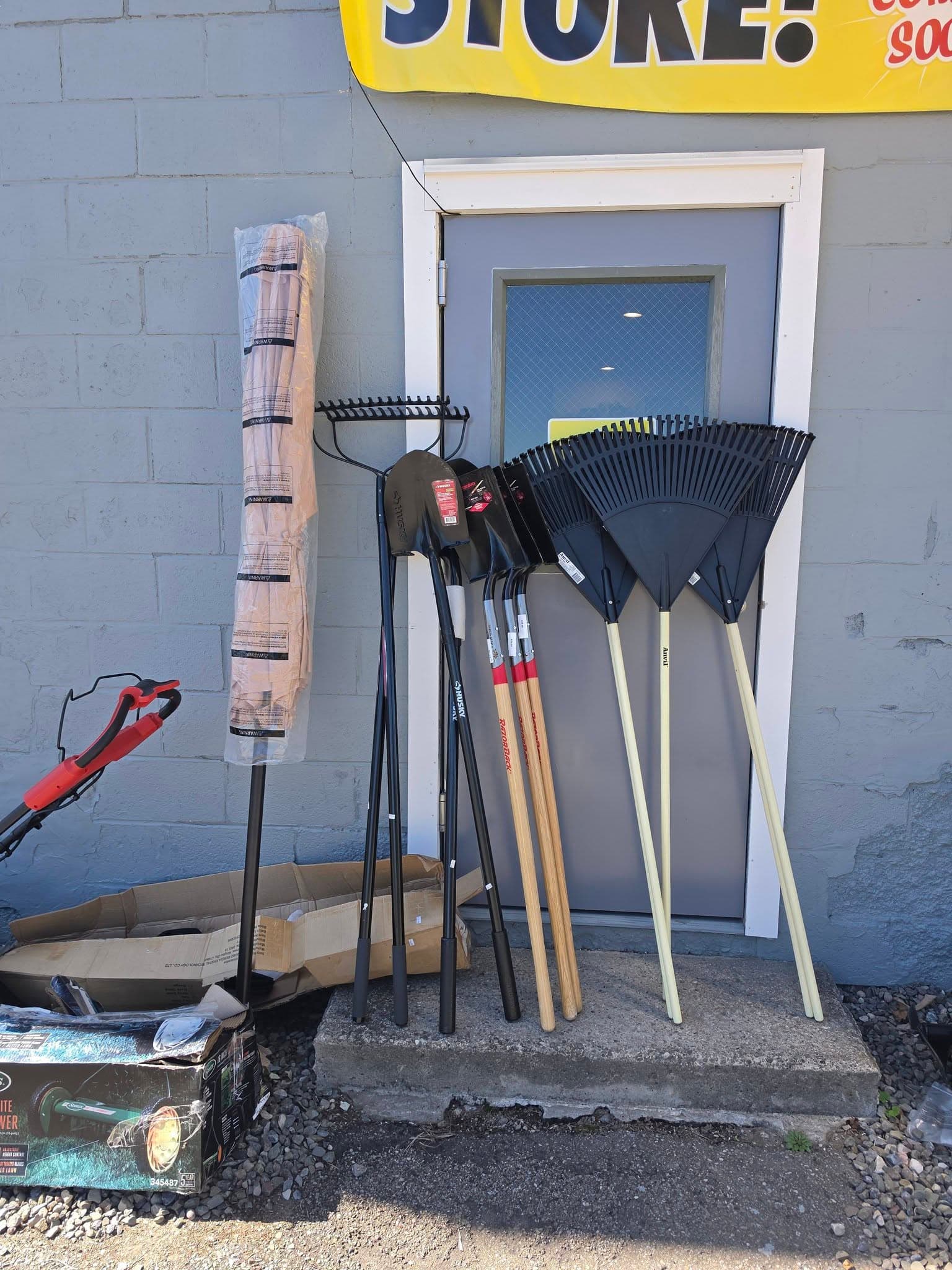 Black shovels, leaf rakes, and a wrapped patio umbrella leaning against a grey store door.