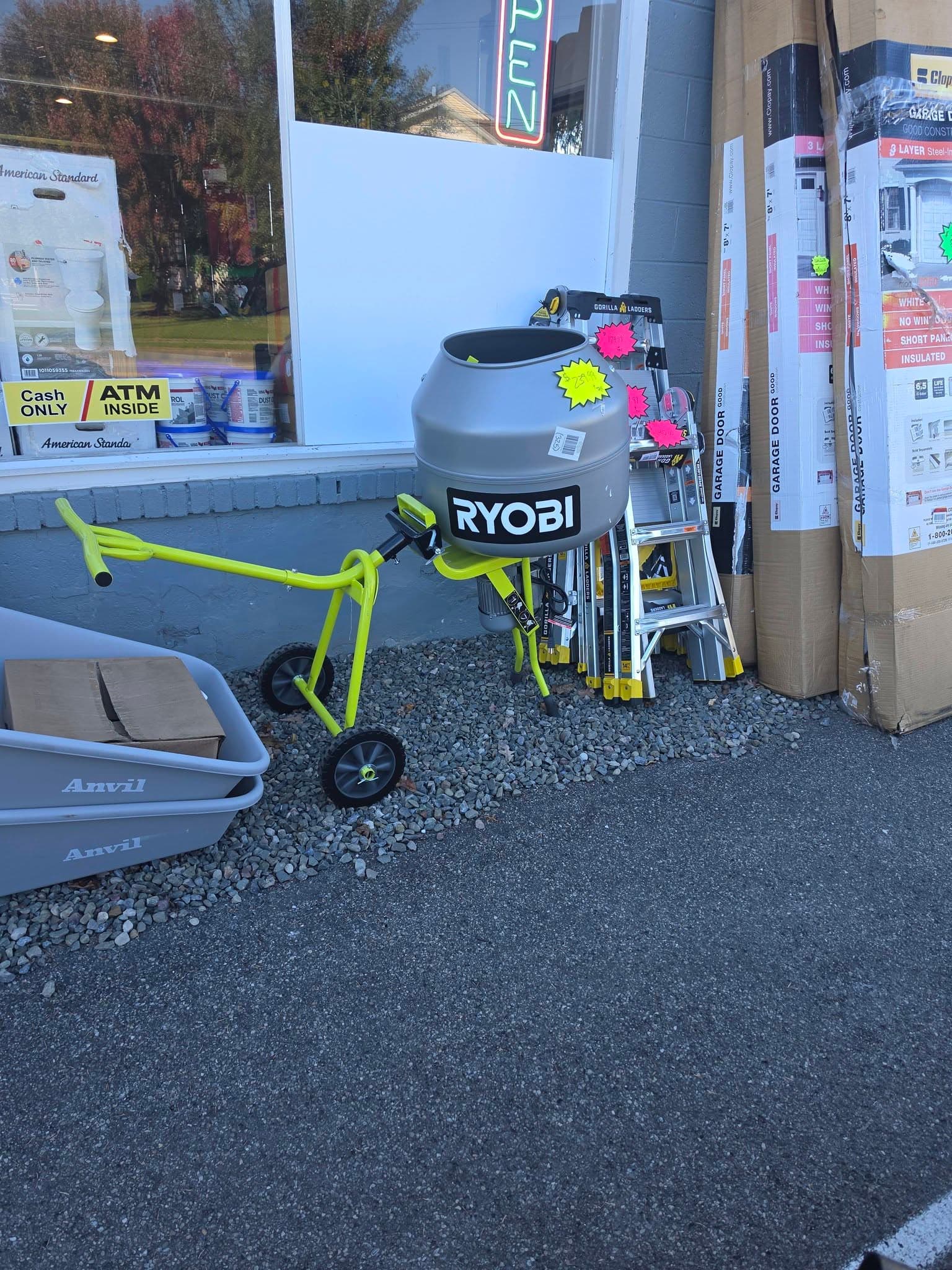 Ryobi cement mixer on a yellow wheeled frame outside a store near ladders and boxes.