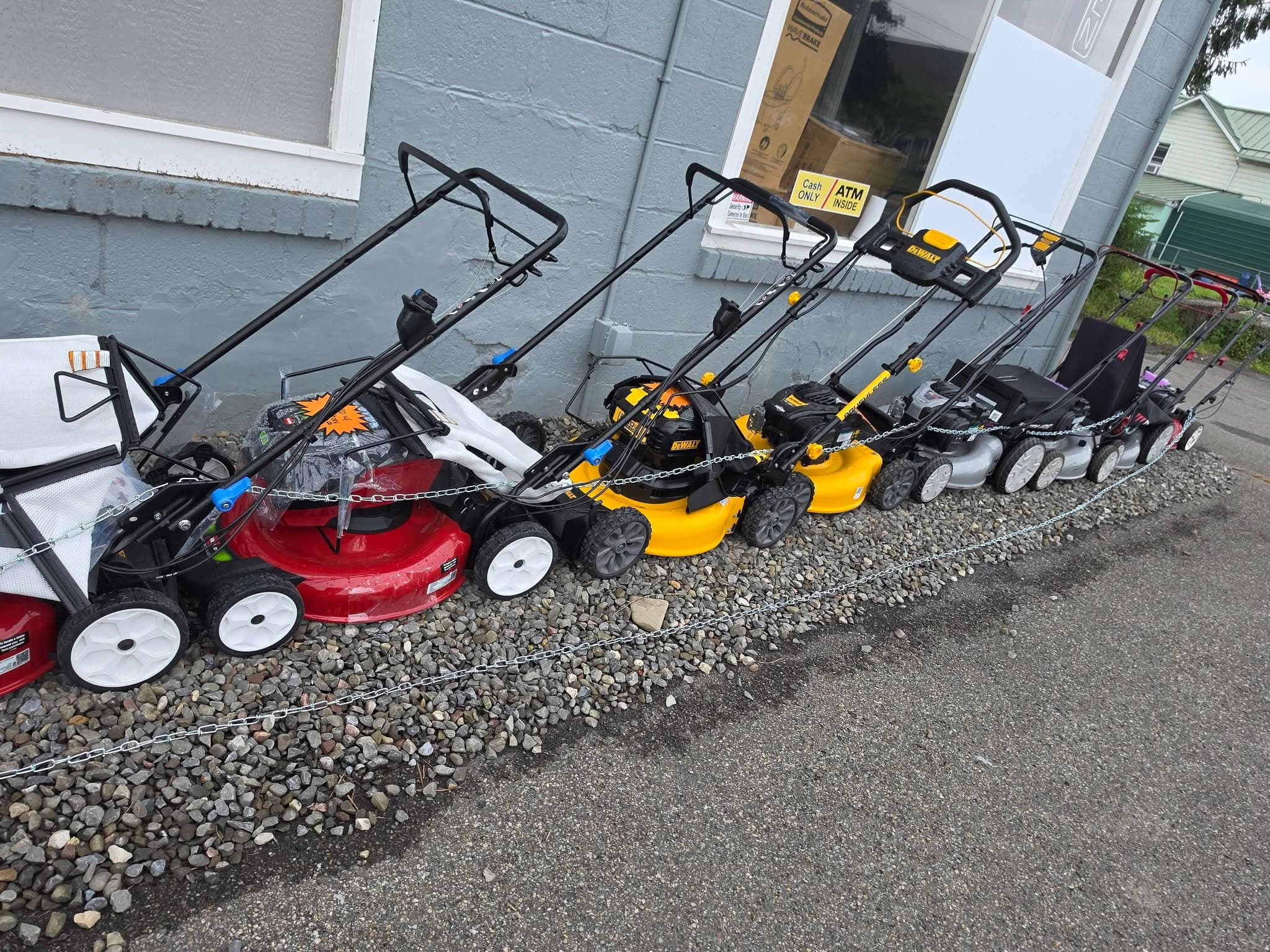 Row of red, yellow, and black lawnmowers chained together on gravel outside a grey building.
